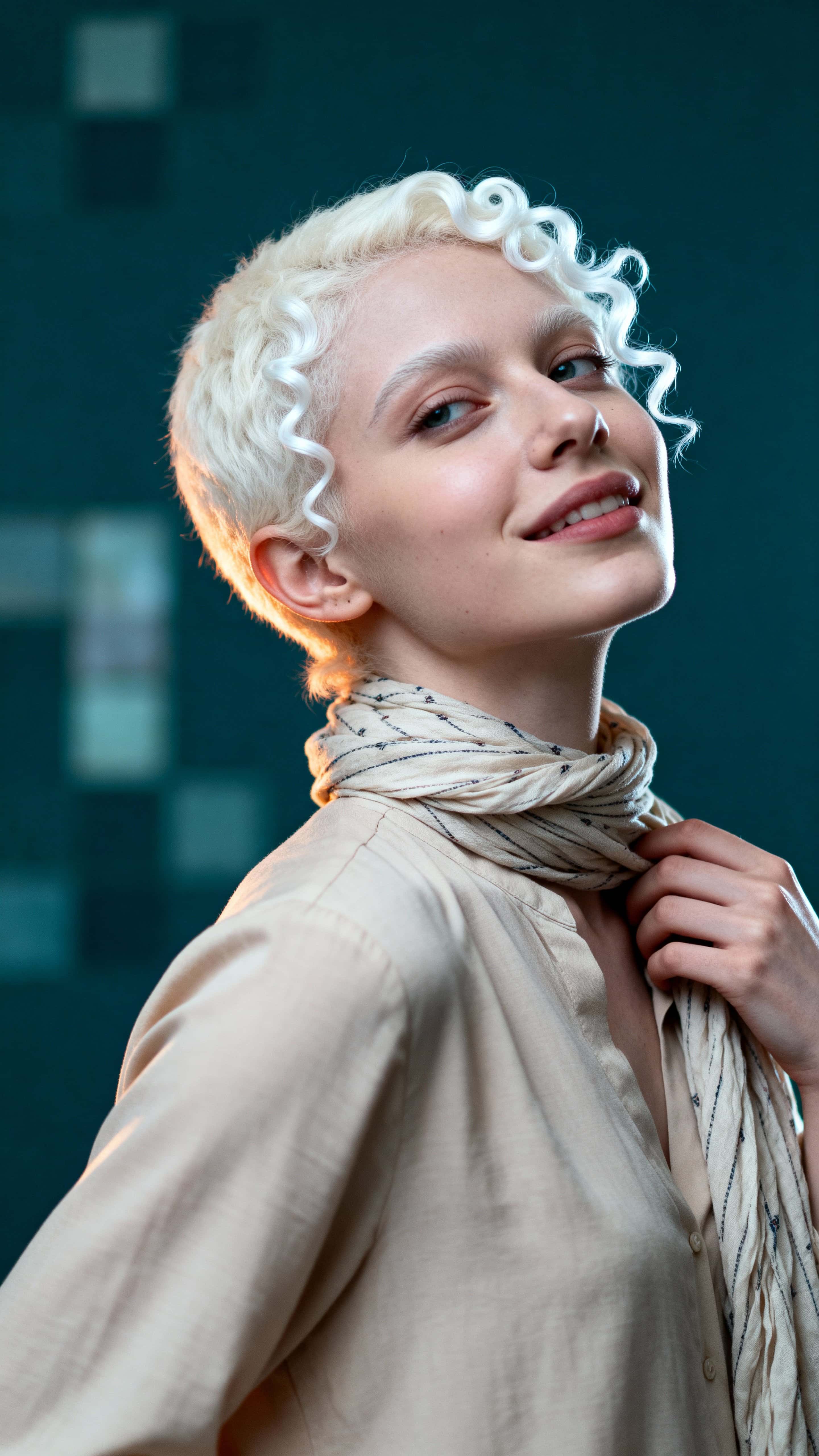 Cinematic shoulders-up portrait of a late-twenties Moroccan woman with albinism against a dark teal, softly mosaic-blurred background, wearing a sand blouse and patterned scarf.