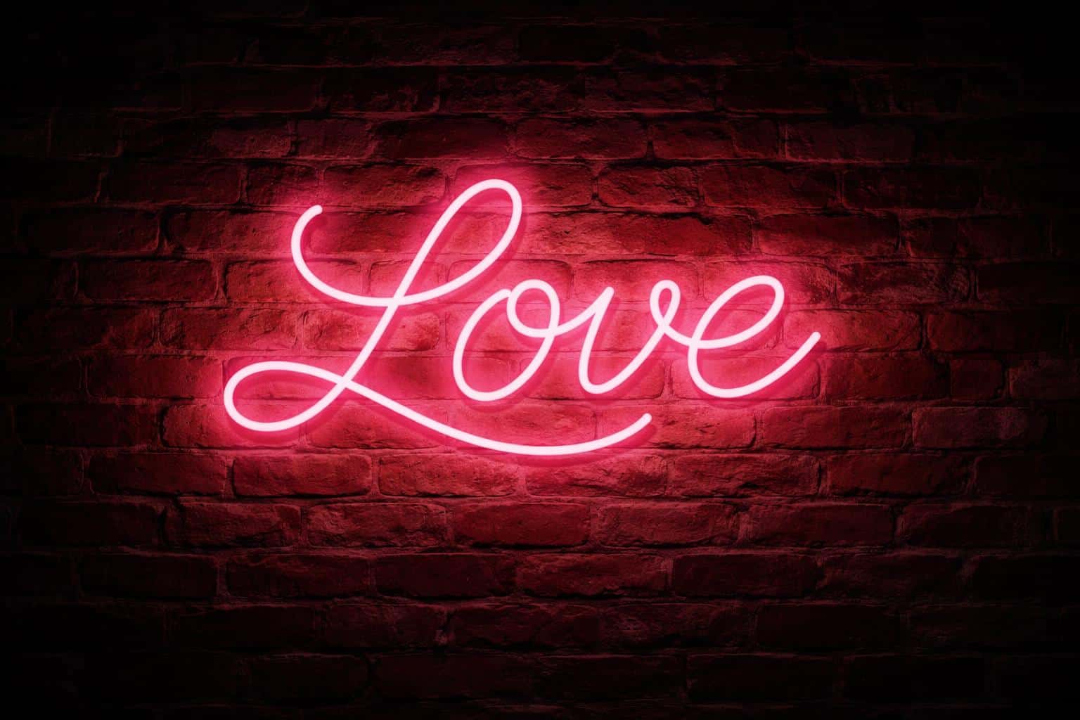 Cinematic 3:2 night photograph of a glowing pink neon 'LOVE' cursive glass sign centered on a weathered red brick wall, casting warm romantic light.