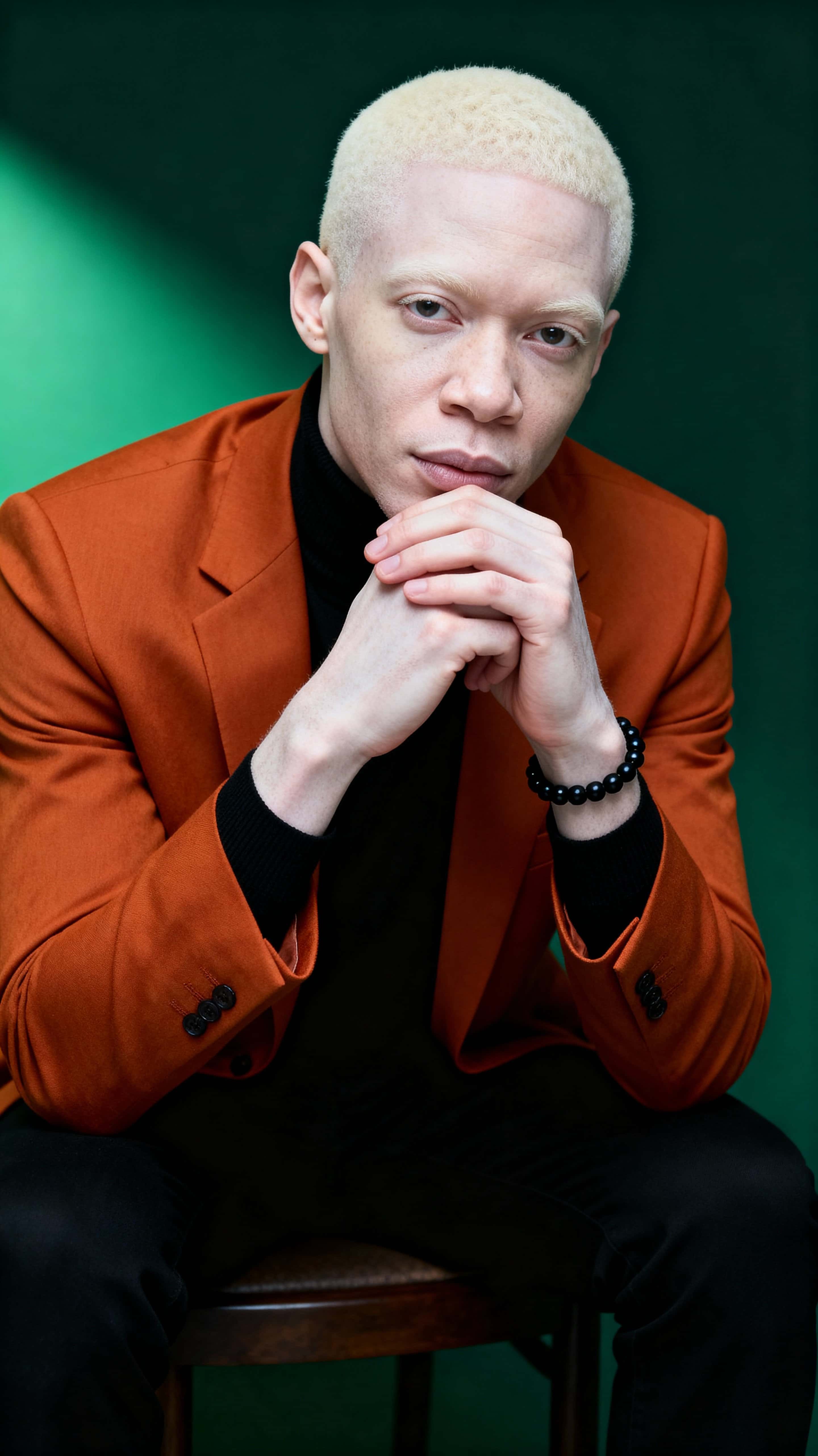 Studio portrait of a 41-year-old Nigerian man with albinism seated mid-thigh up, leaning forward with steepled hands, wearing a burnt orange blazer and black turtleneck against a deep charcoal-green background.