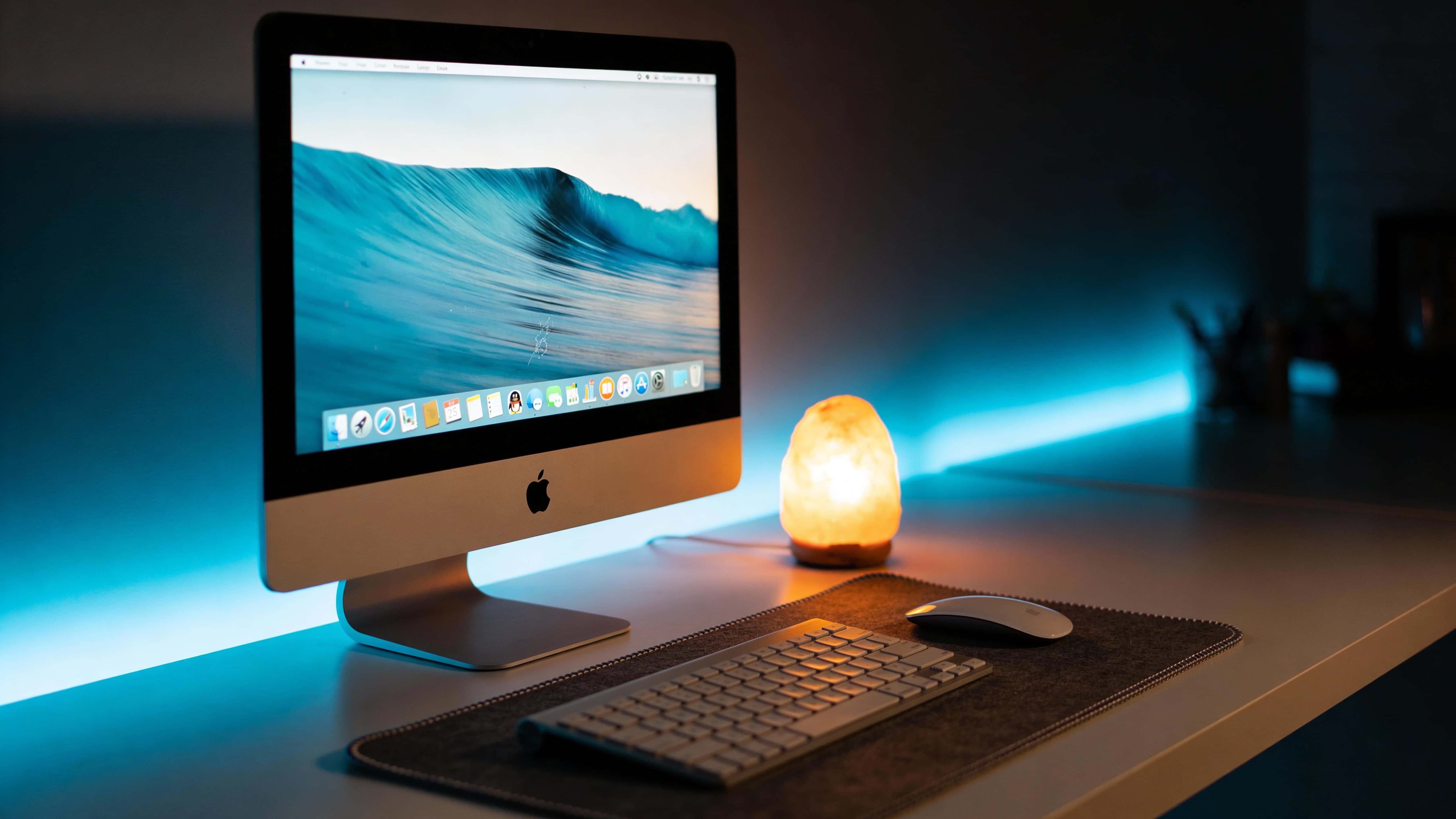 Night desk scene photographed as an interior photography composition with cool monitor glow, warm accent lamp, and explicit 16:9 framing for wallpaper or background use.