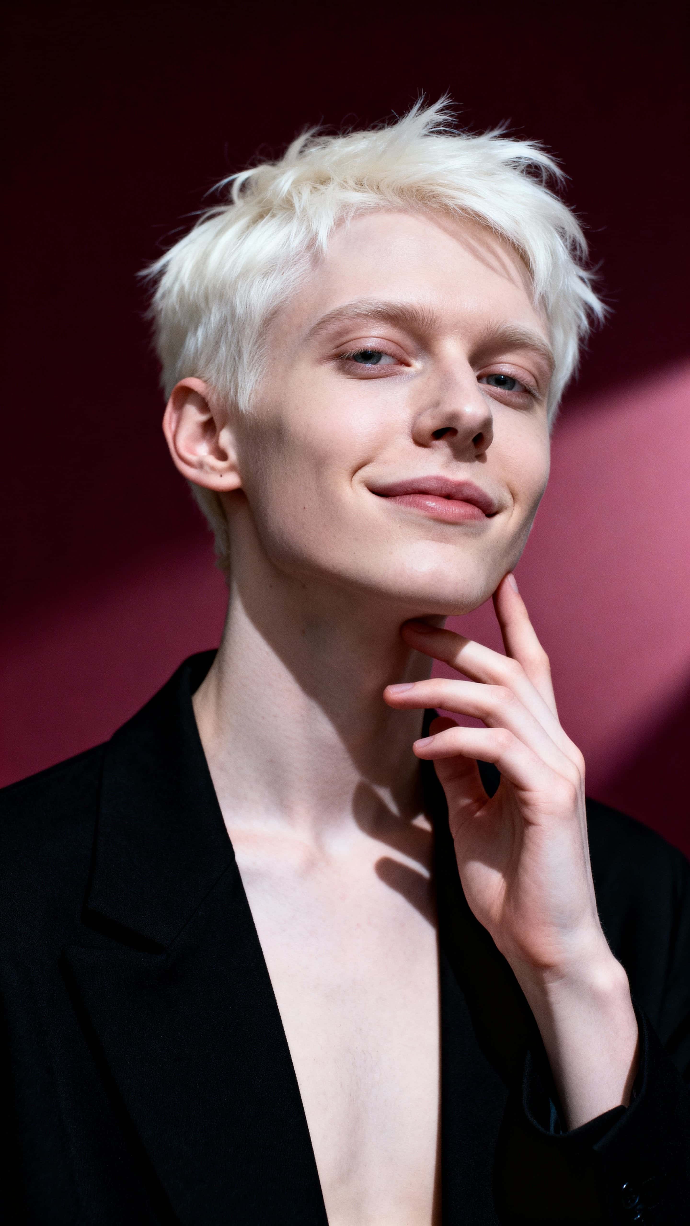 Introspective studio portrait of a late-twenties Eastern European non-binary person with albinism, short white hair, minimalist black jacket, wine-red backdrop.