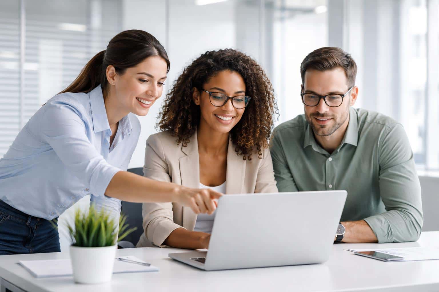 Lifestyle photography of three diverse professionals collaborating over a laptop in a bright, modern office space with glass partitions and high-key light.