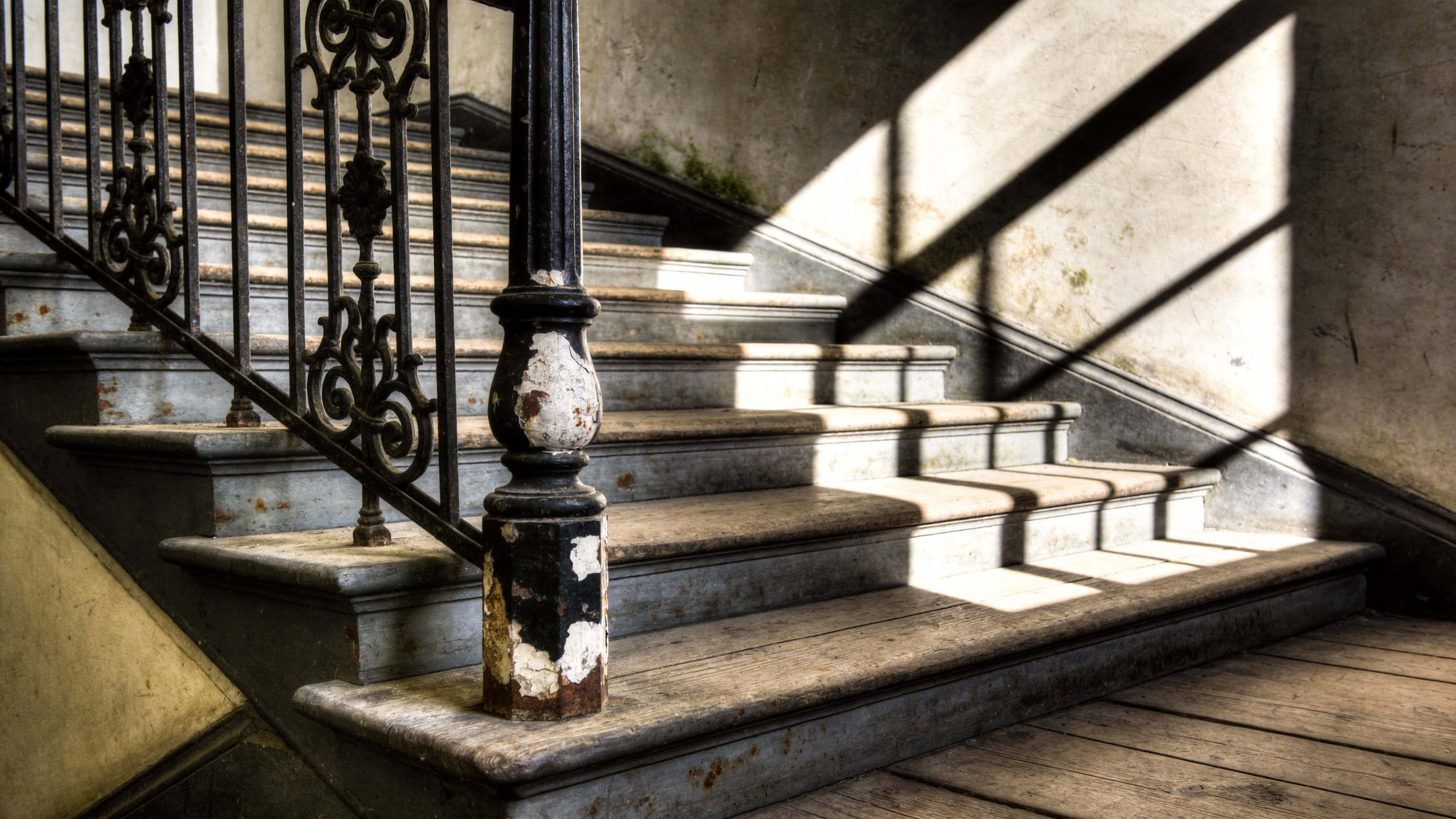 A 16:9 architectural photograph of an old staircase with peeling paint, worn wooden treads and dramatic side-lit shadows, composed for wallpaper or poster use.