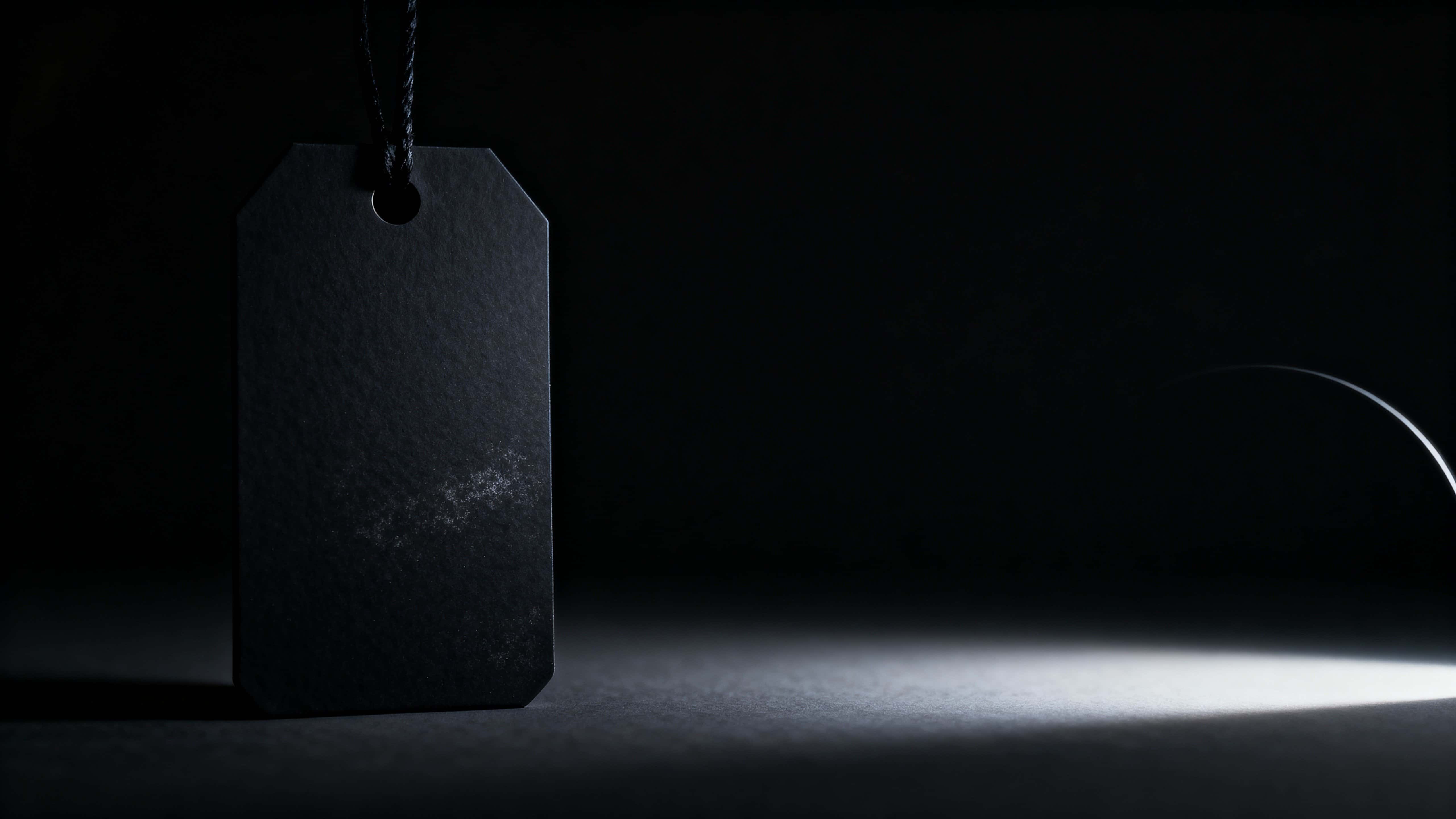 A single oversized matte-black price tag floats mid-left in a 16:9 frame with subtle rim lighting, powdery rubber texture, soft edge haze, and expansive near-black negative space.