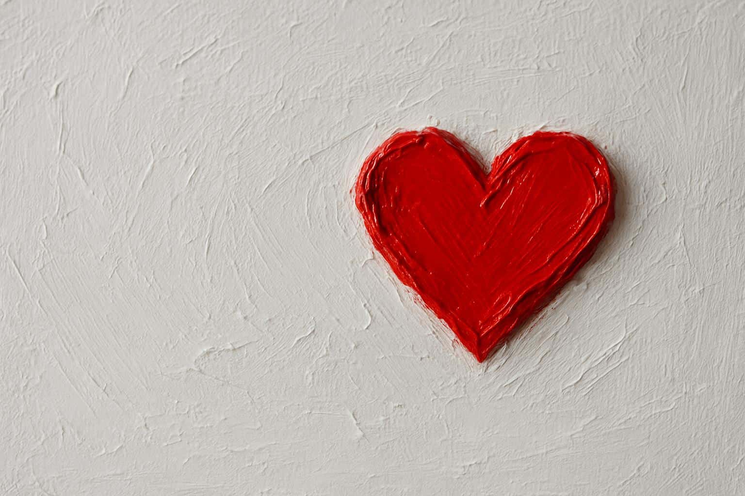 A small vivid red painted heart rendered in thick impasto sits slightly above center on a white textured canvas, with generous negative space to the left and top, soft upper-left light, glossy wet edges on the red paint, and a macro still-life focus that preserves tactile palette-knife strokes and sculptural off-white paste within a 3:2 frame.