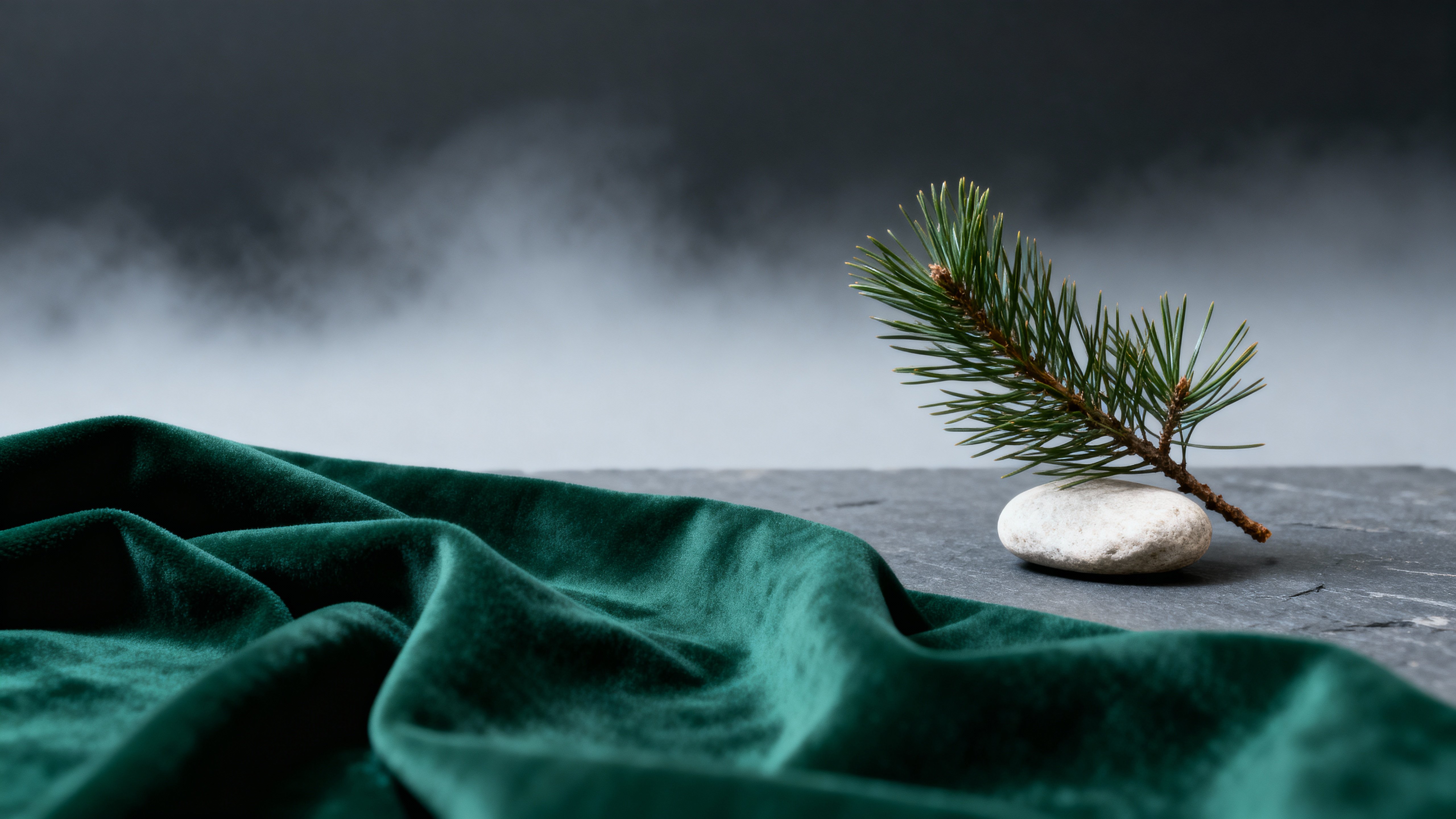 Pine sprig on deep-green textile folds in the foreground against a cool slate fog gradient, balanced by a pale stone; sprig and stone are crisply focused while folds dissolve softly, leaving open upper third for typography.
