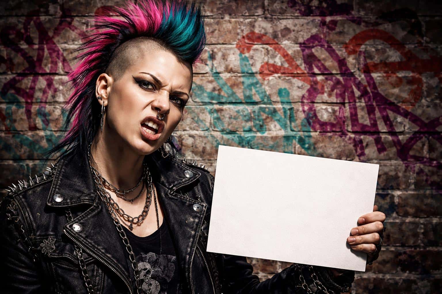 A defiant punk rocker holds a blank placard against a graffiti-splattered brick wall, lit by harsh side light that sculpts deep shadows and sharp highlights for a poster-ready portrait.