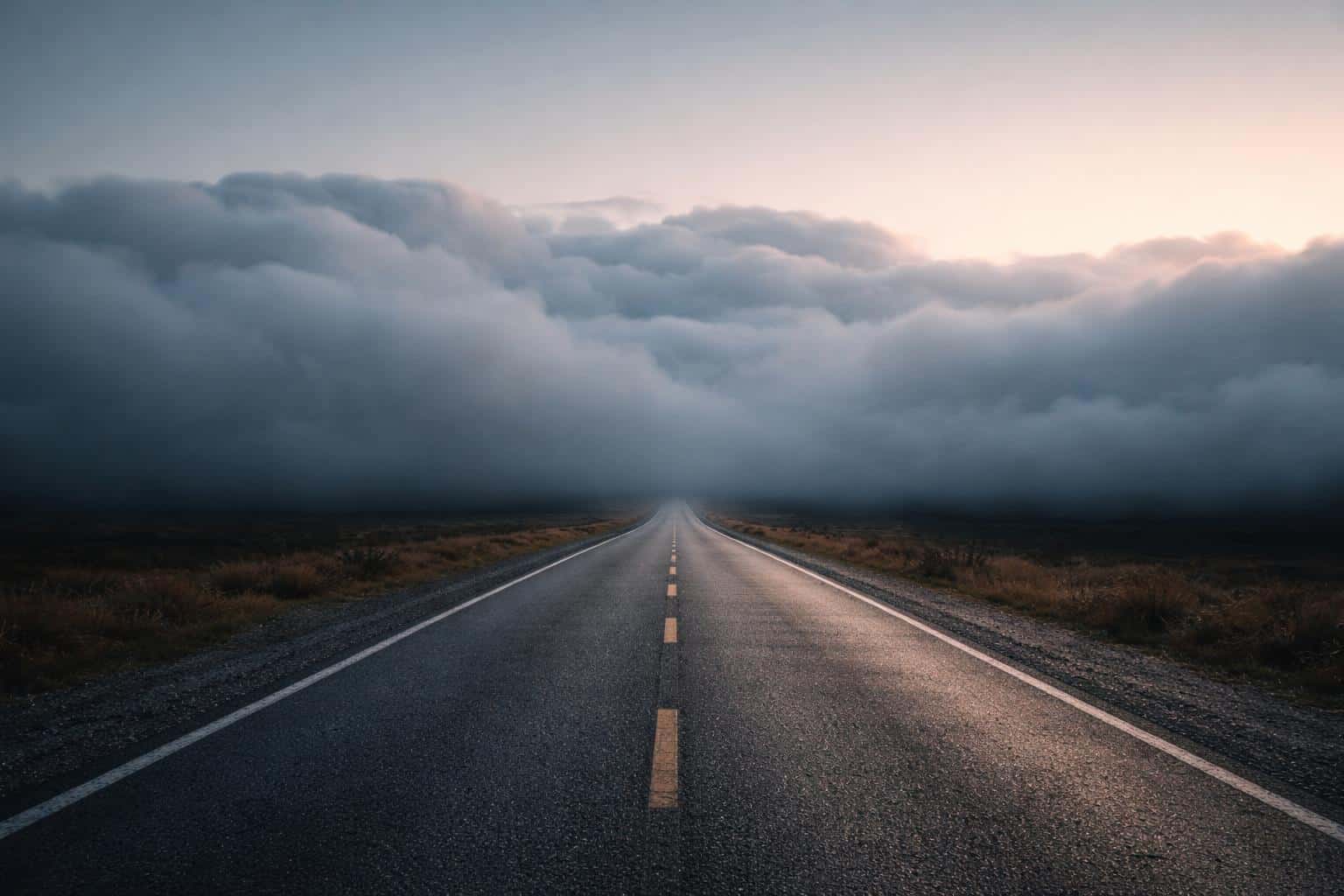 A wide 3:2 landscape photograph of a lone road that disappears into low clouds at the horizon, with moody light and minimalist composition for wallpaper use.