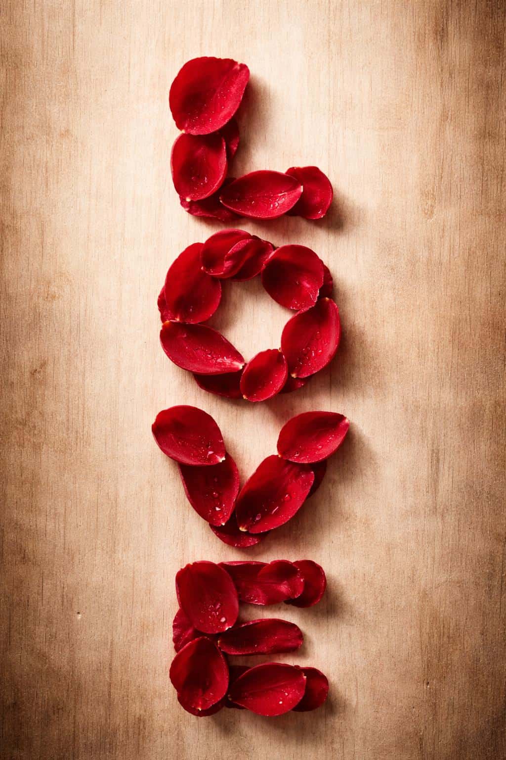 Cinematic macro portrait of the word LOVE formed from fresh crimson rose petals stacked vertically on a rustic light-wood background with soft, minimalist romance.