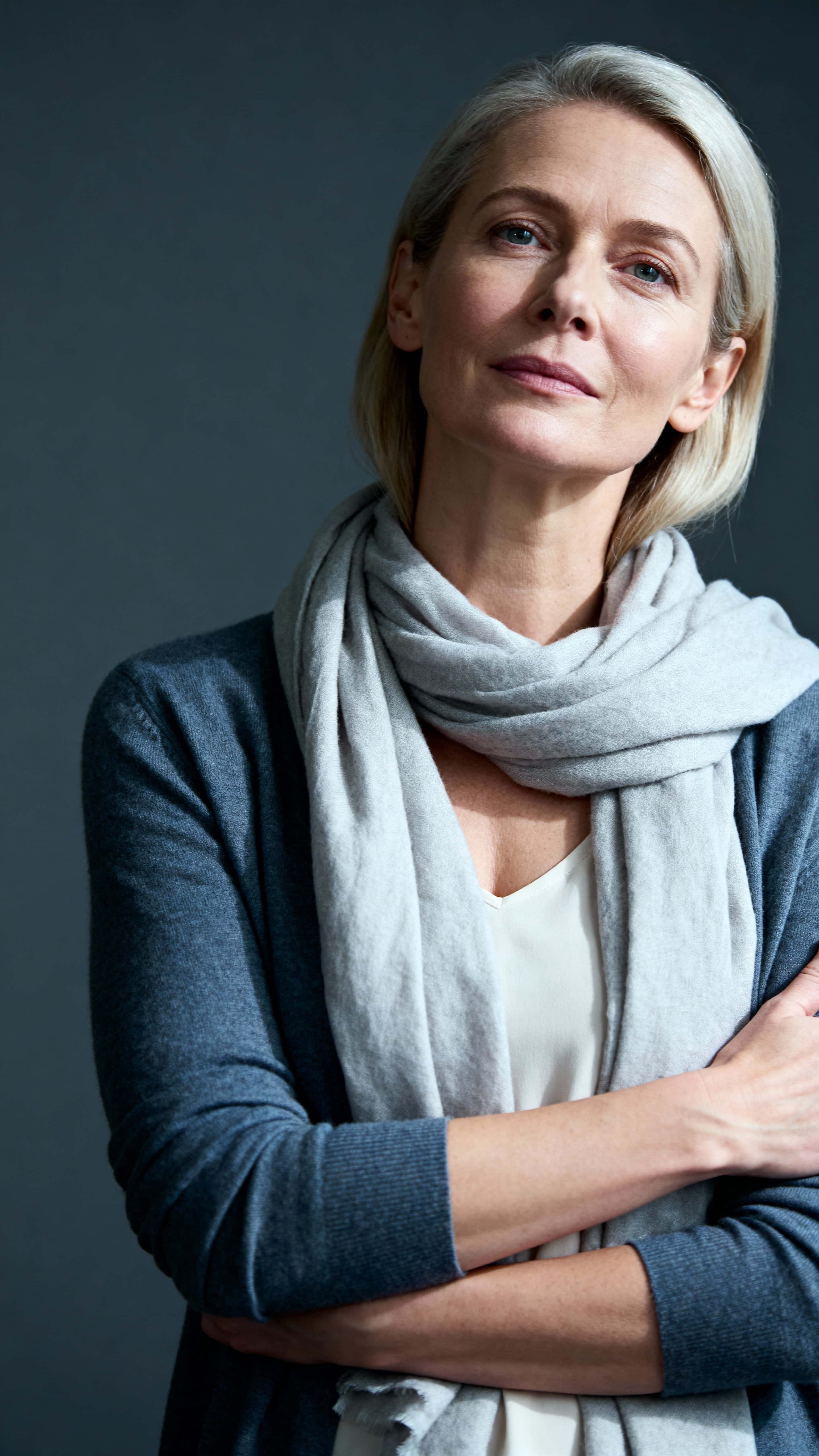 A gentle studio portrait of a 45-year-old Scandinavian woman with alabaster skin and ash-blond hair, wearing a slate-blue cardigan and light gray scarf against a dark steel-gray background.