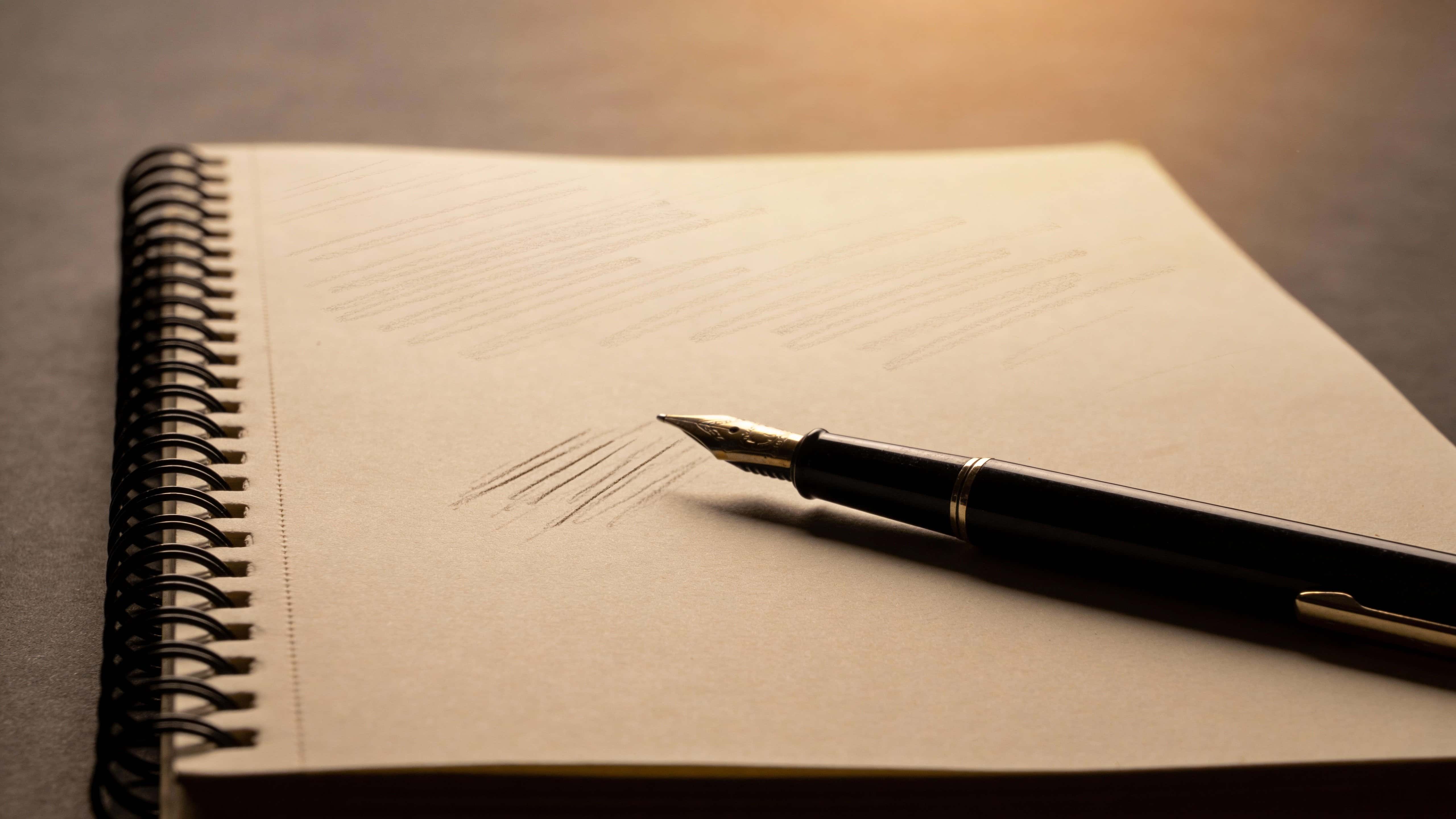 Close-up product photograph of a sketchbook and pen in a 16:9 layout, tightly composed for wallpaper or poster use with warm paper tones and soft shadows.