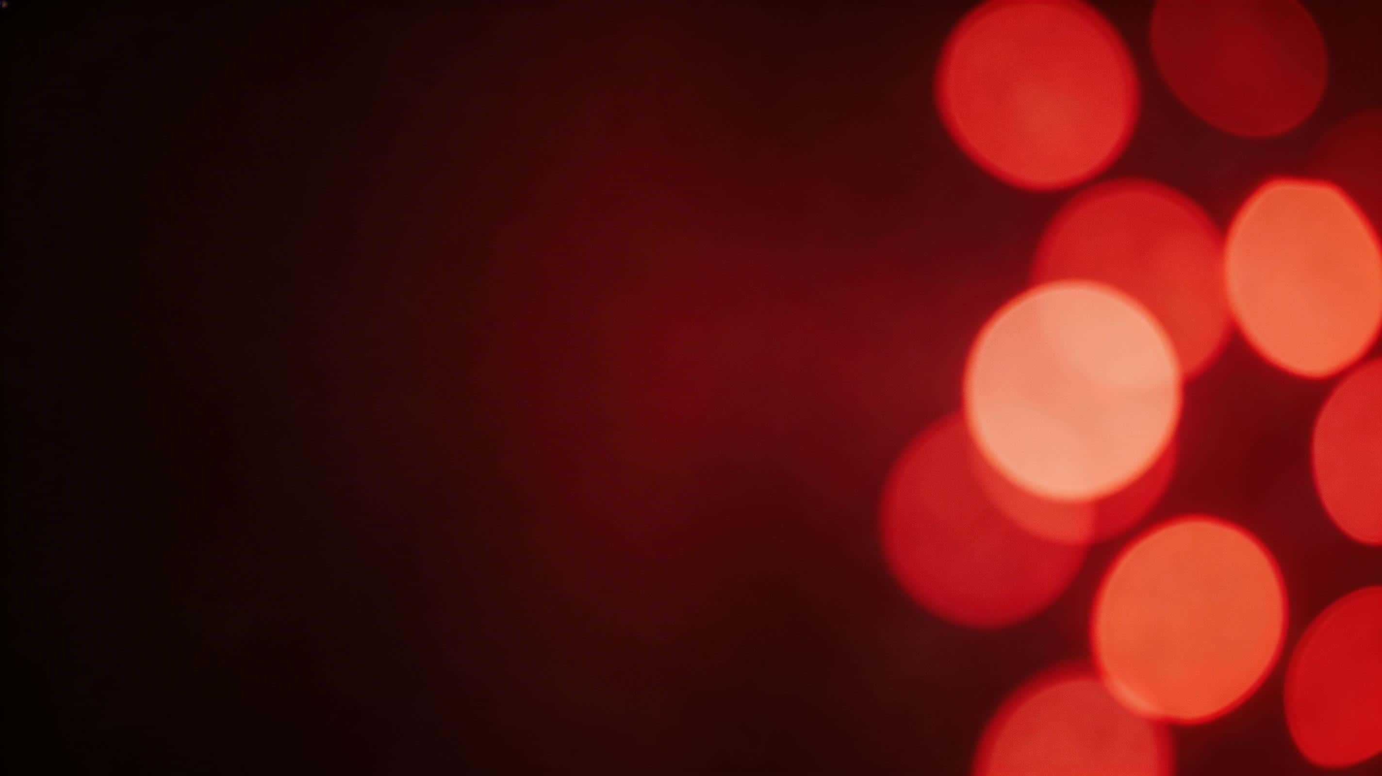 Cinematic 16:9 editorial photograph of a soft red bokeh light wall with large defocused orbs clustered to one side, a clean dark gradient negative-space zone, and subtle haze.