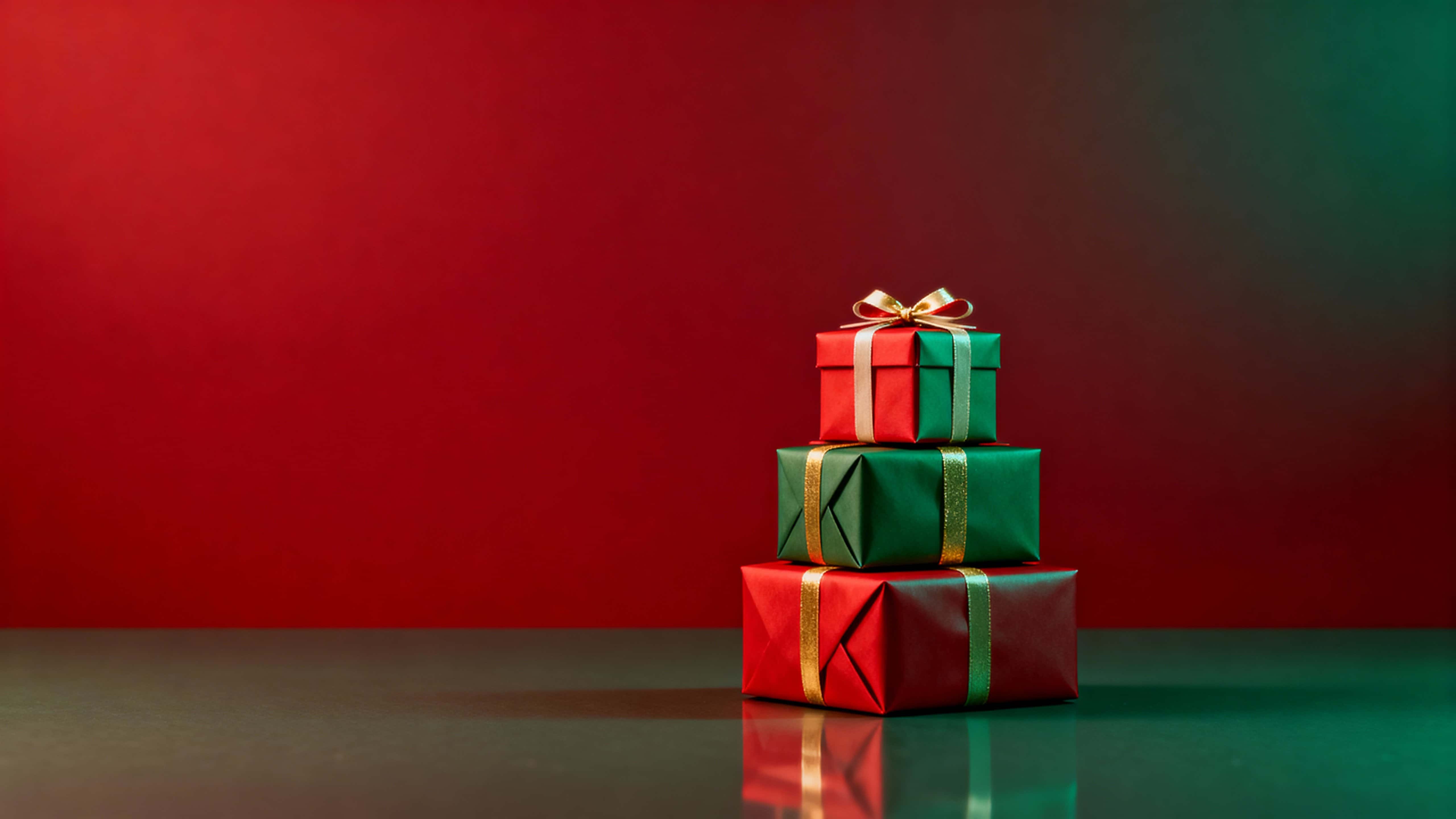 Horizontal Christmas scene with a small tower of red, green and gold wrapped gift boxes stacked slightly right of center on a subtle reflective surface, leaving calm copy space above and left.