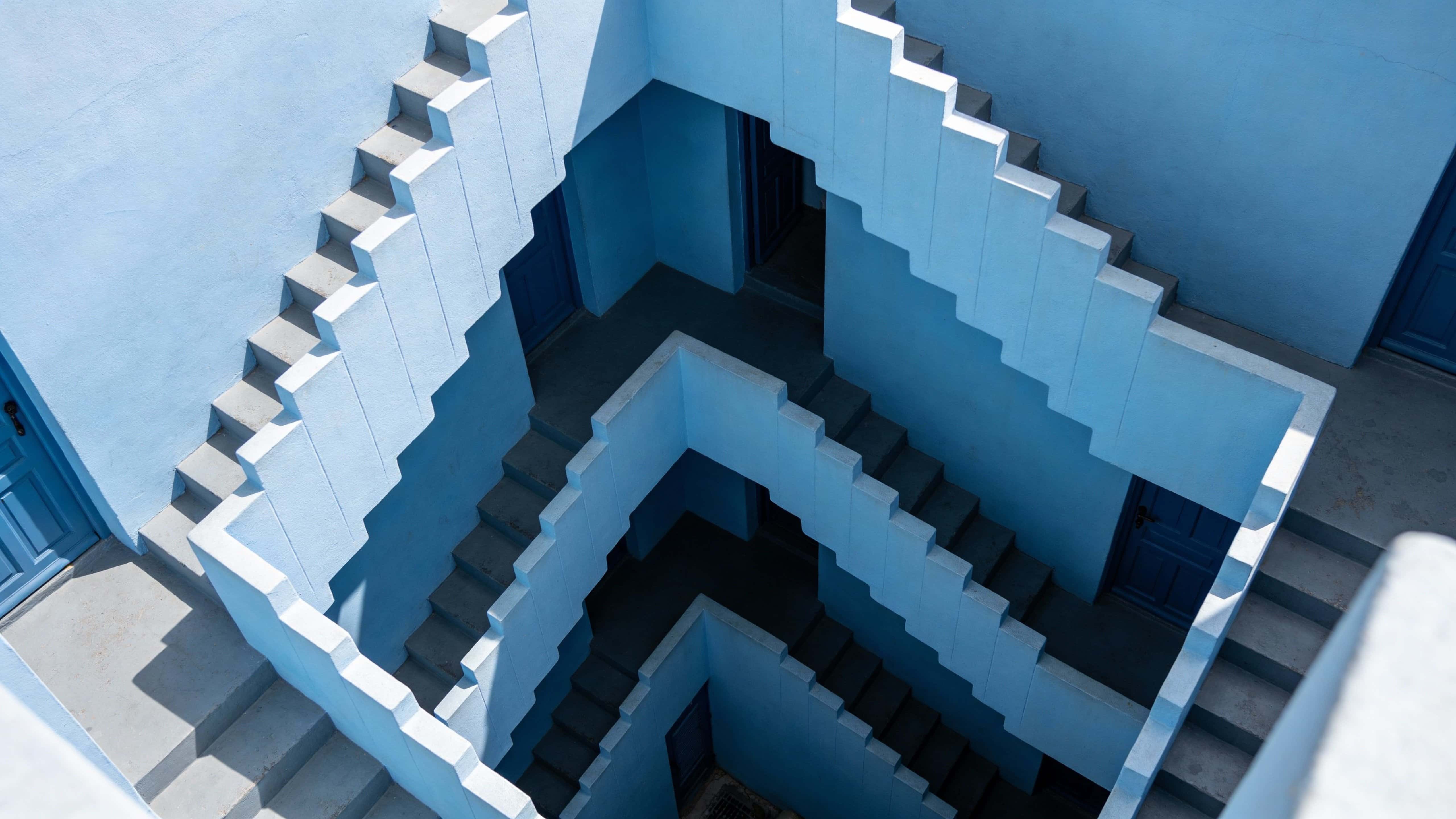 Top-down angled 16:9 view of interconnected staircases and corridors in Mediterranean-inspired minimalist brutalist architecture, painted in monochromatic blue with soft daylight and subtle shadows.
