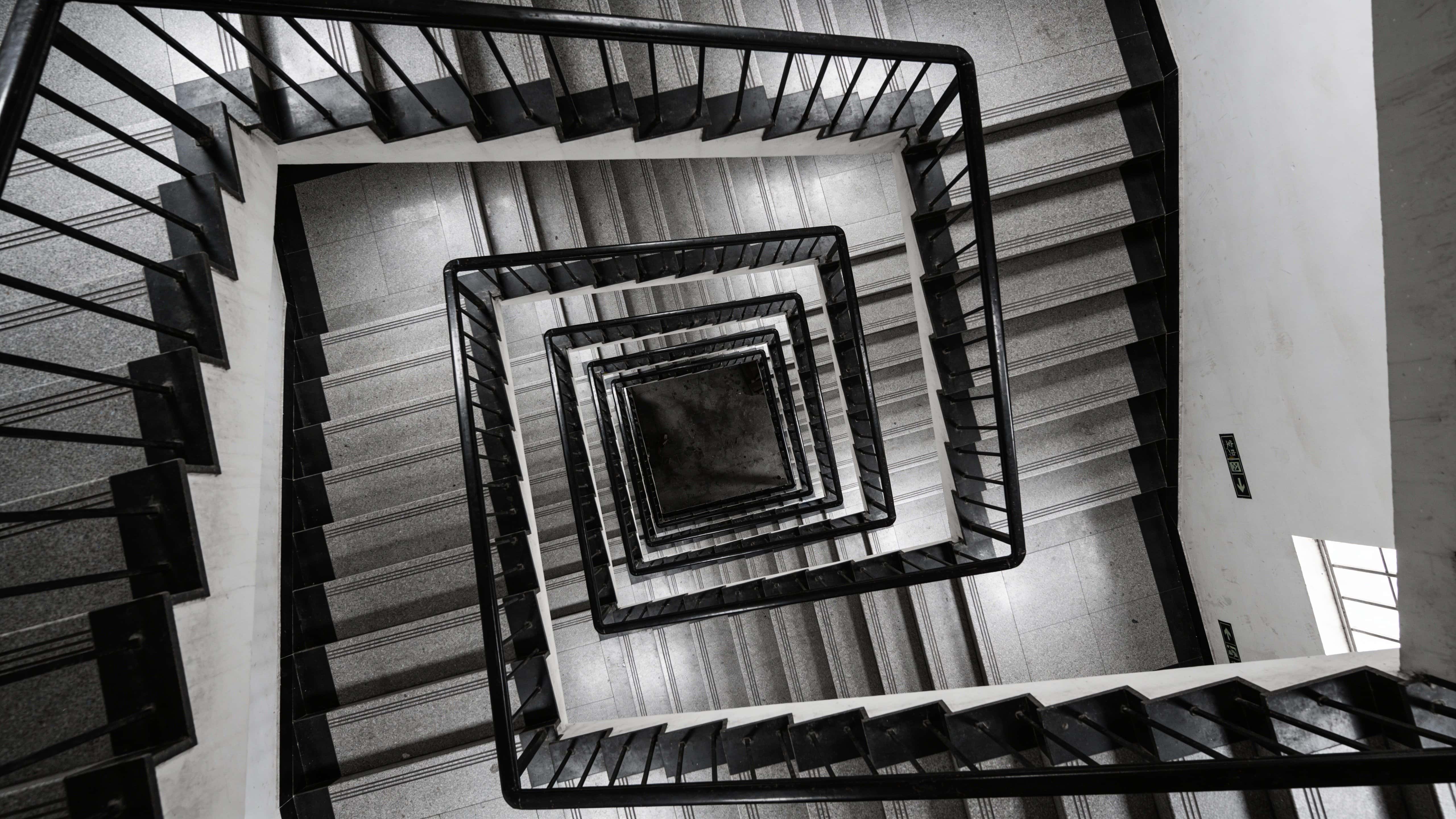 An intense architectural maze of tightly spaced staircases and narrowed corridors, concrete walls and metal railings creating repeating angular patterns and claustrophobic depth across a 16:9 horizontal frame.