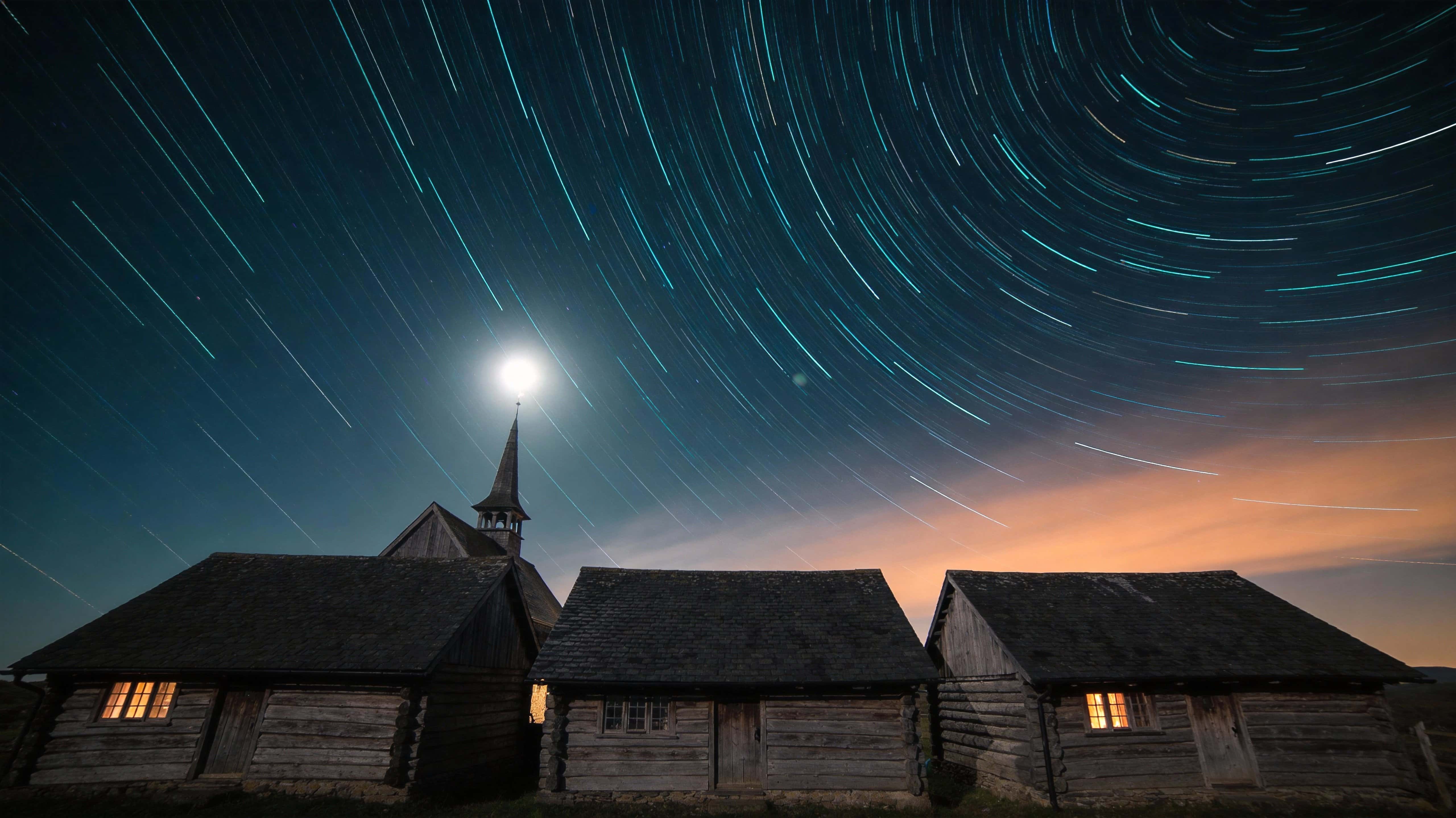 Long-exposure landscape photograph of star trails arcing over a quiet rural village at night, composed for a 16:9 wallpaper-style frame with cinematic crop.