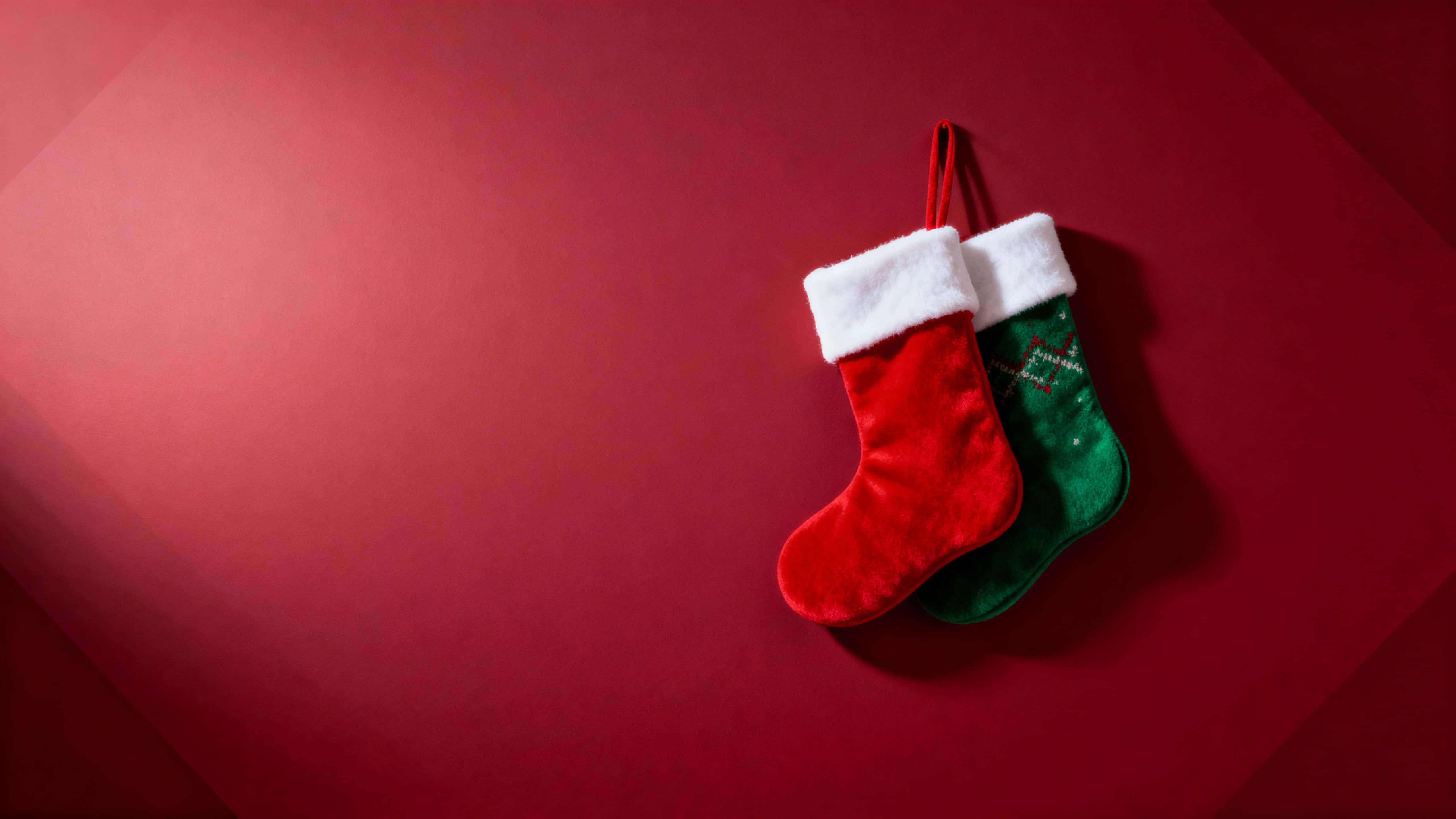 A 16:9 horizontal scene of two cozy fabric stockings—one red with white cuffs, one patterned green—hung at the right with a deep crimson gradient background and calm left-side space for copy.