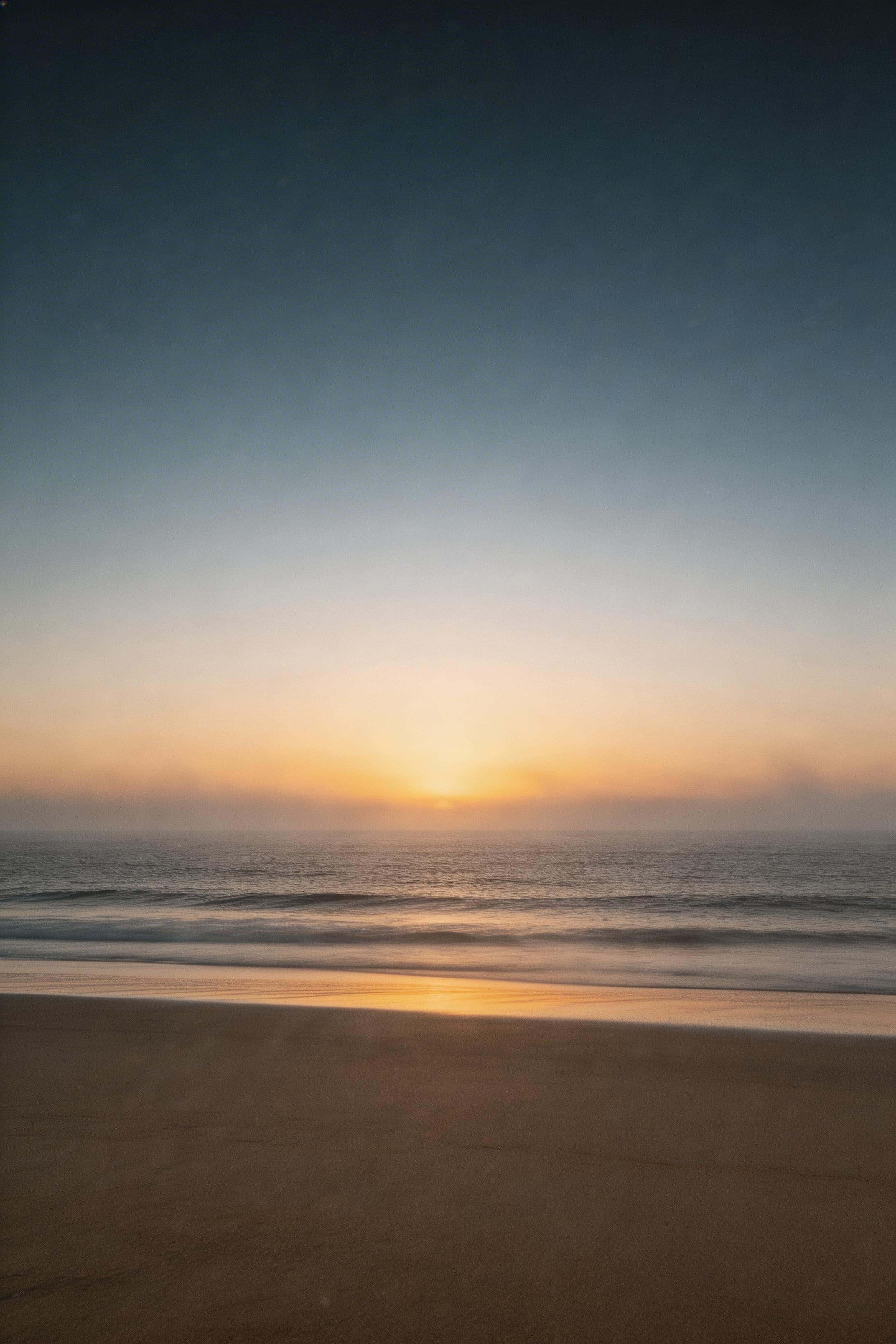 Cinematic editorial 2:3 vertical wallpaper of a minimal sunrise beach with the brightest glow near the lower horizon, upper sky fading into blue-hour haze, subtle mist and fine film grain for realistic texture.