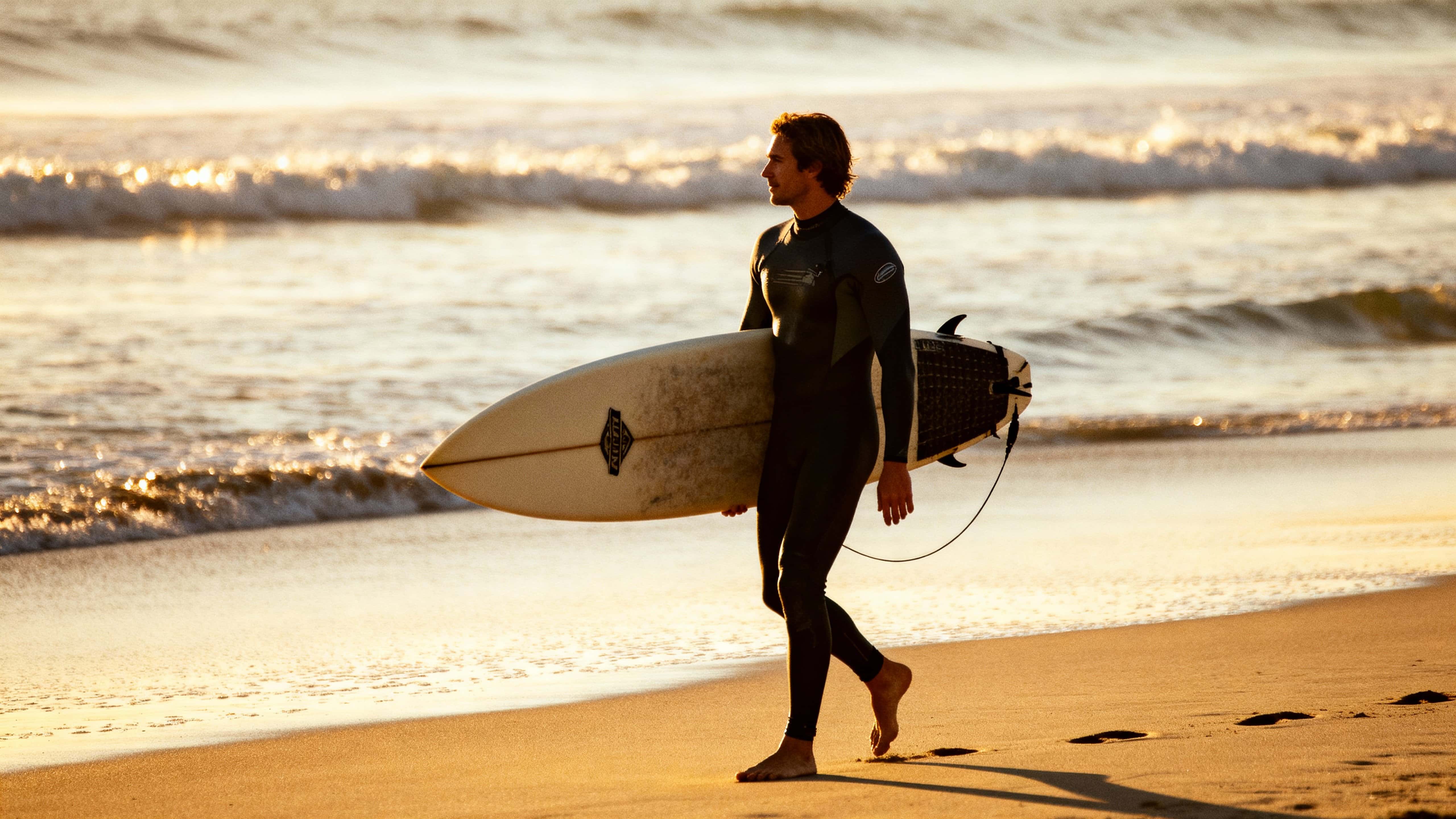 A lone surfer walks along Malibu beach at golden morning light, carrying a board as waves shimmer and footprints trail in the sand, framed wide for a cinematic 16:9 shot.