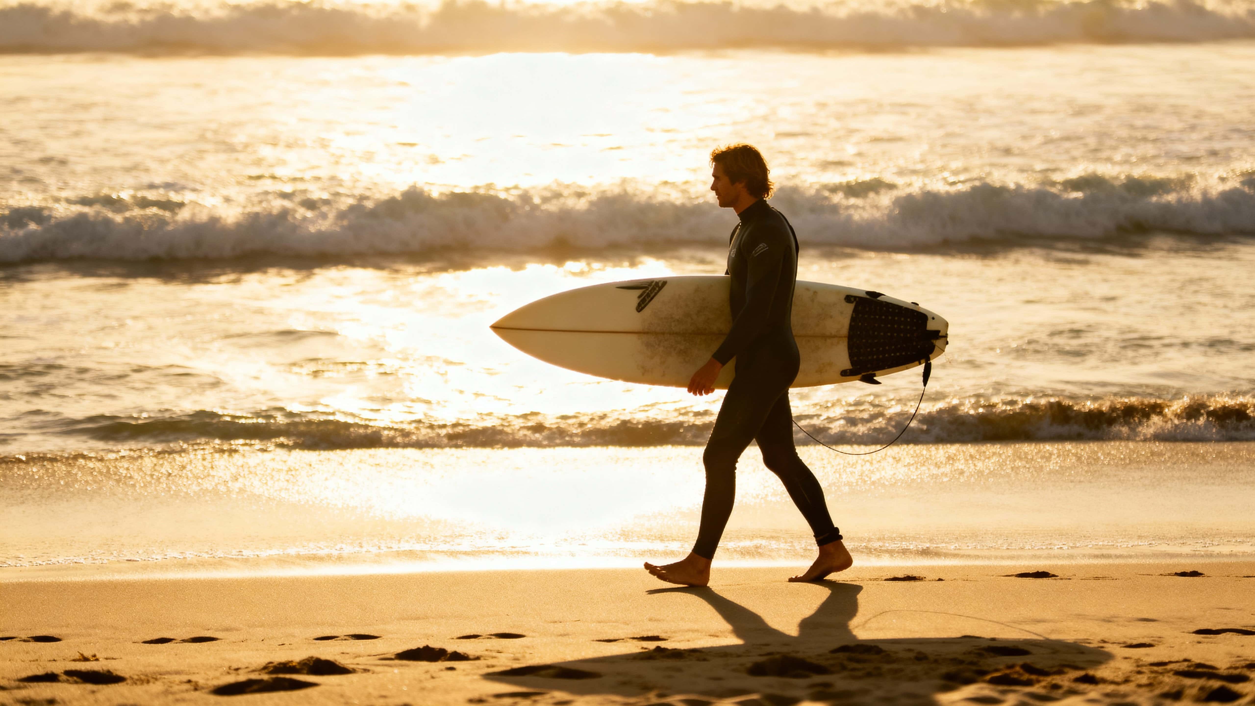 A lone surfer walks along Malibu beach at golden morning light, carrying a board as waves shimmer and footprints trail in the sand, framed wide for a cinematic 16:9 shot.