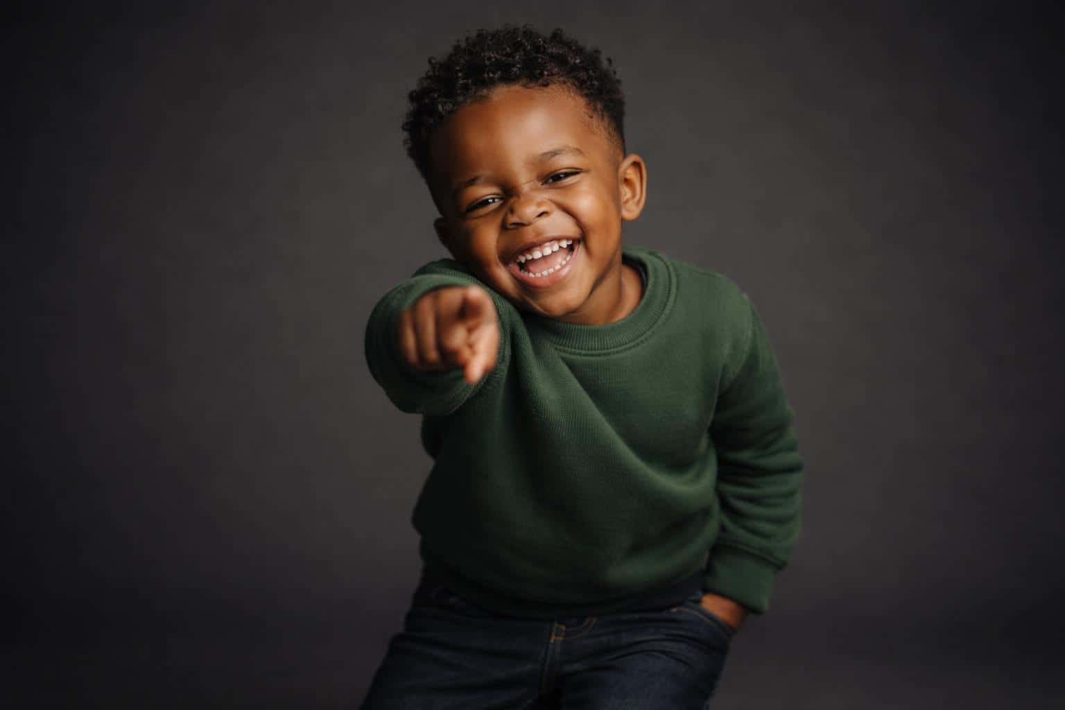 Cinematic 3:2 editorial studio portrait of a grinning three-year-old Black boy with tight curls leaning forward and pointing at the lens in a forest-green sweater on a seamless deep-charcoal backdrop with warm amber highlights.