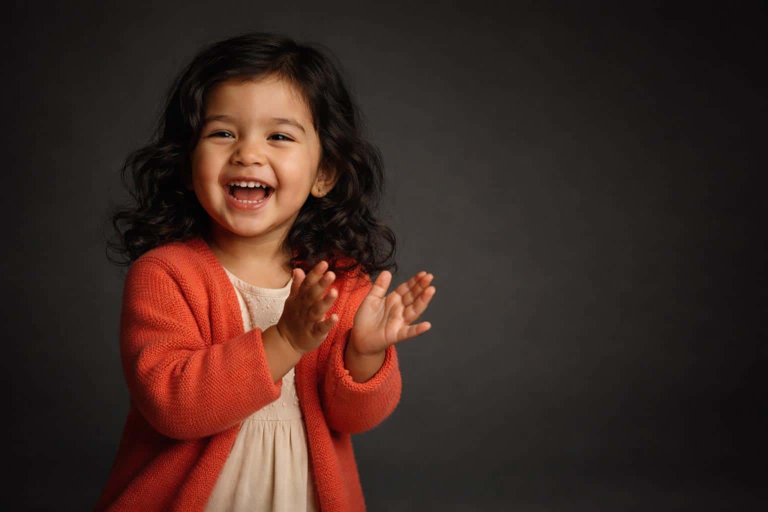 Cinematic 3:2 editorial studio portrait of a laughing three-year-old Latina girl in a coral cardigan and cream dress on a deep charcoal seamless backdrop, hands mid-clap with soft wrapped light, subtle rim, shallow depth and gentle film grain.