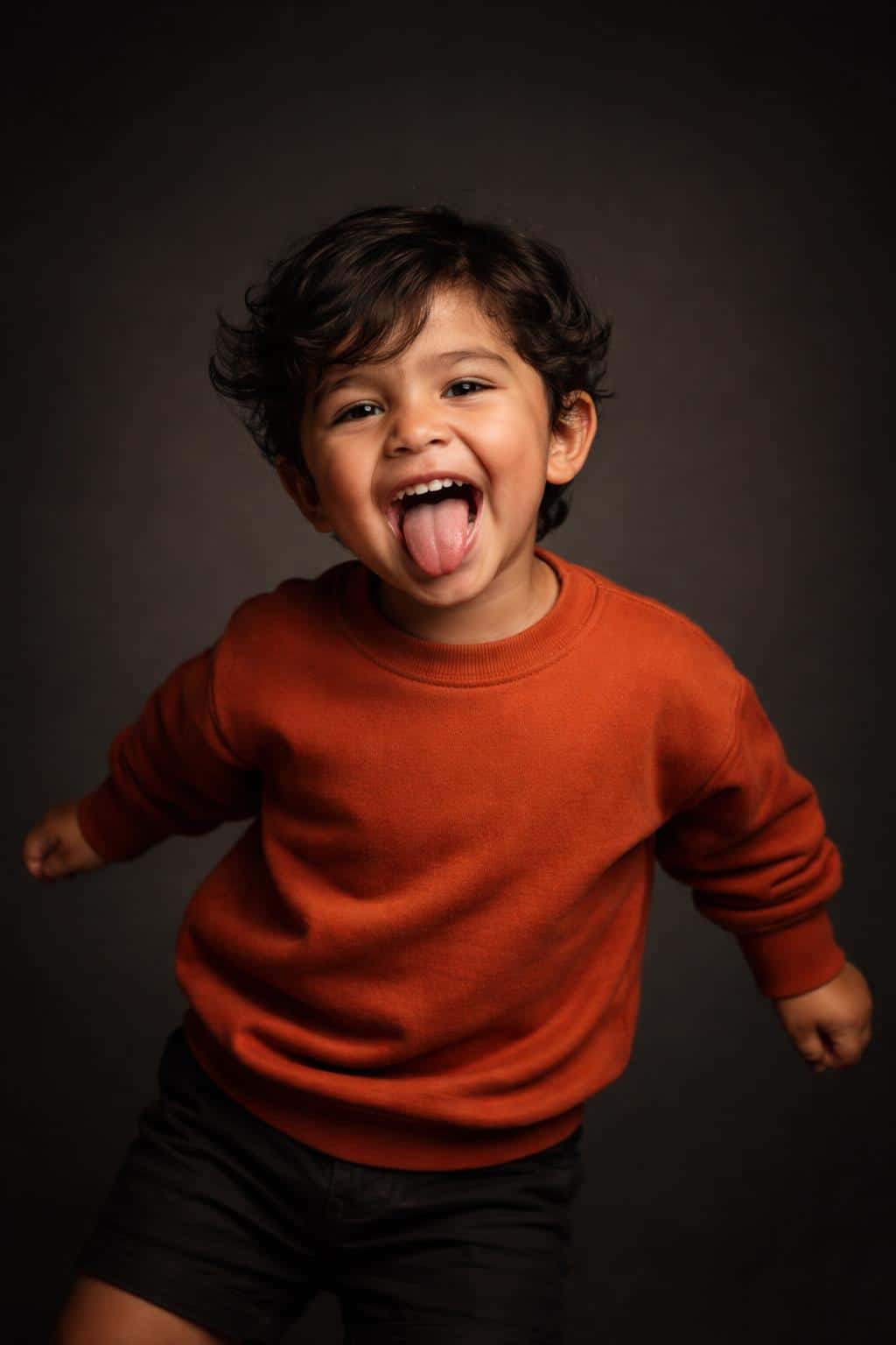 Cinematic 2:3 studio portrait of a laughing three-year-old Latino boy in a burnt-orange sweatshirt against a minimal dark gradient with warm amber highlights and subtle grain.