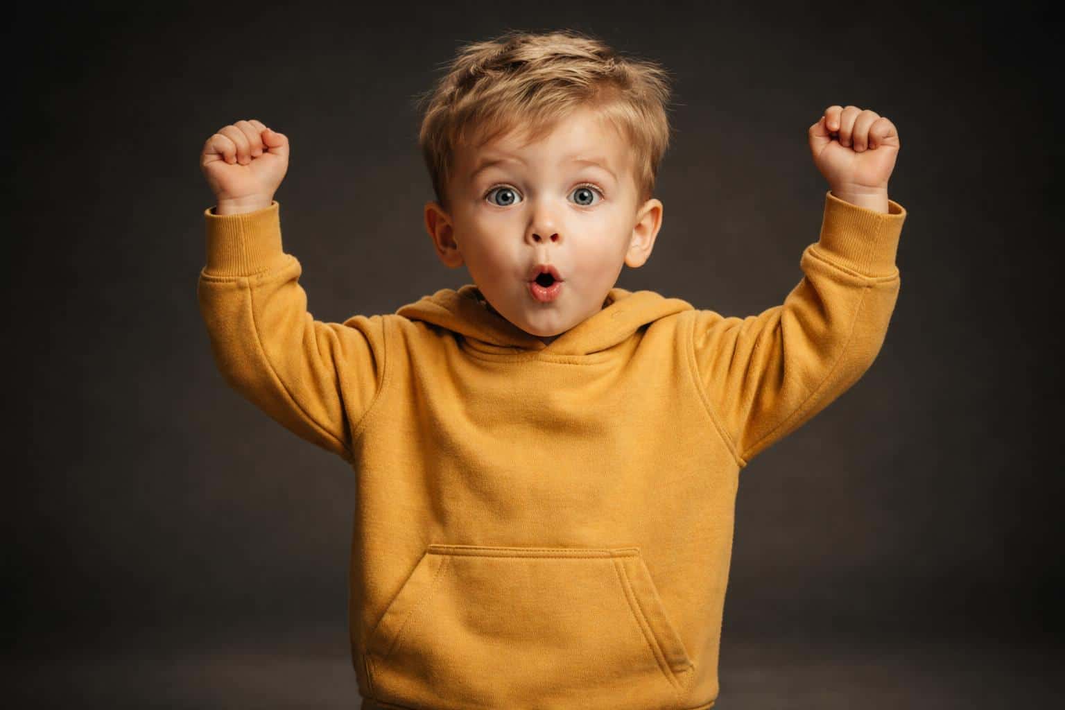 Wide 3:2 cinematic studio portrait of a two-and-a-half-year-old fair-skinned sandy-blonde boy in a mustard hoodie making an O-shaped mouth with arms raised against deep charcoal backdrop.