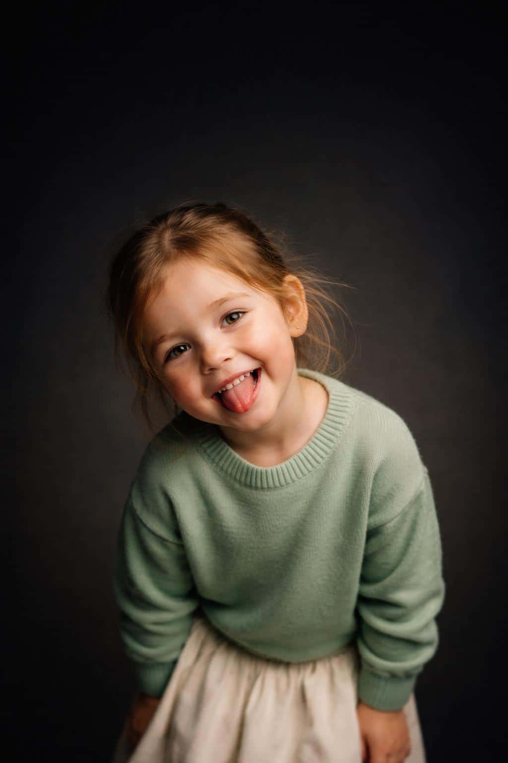 Cinematic 2:3 vertical studio portrait of a three-year-old fair, freckled toddler girl in a mint sweater and cream skirt, playful tongue-out grin, lower-middle framing with a minimal dark-gradient background.