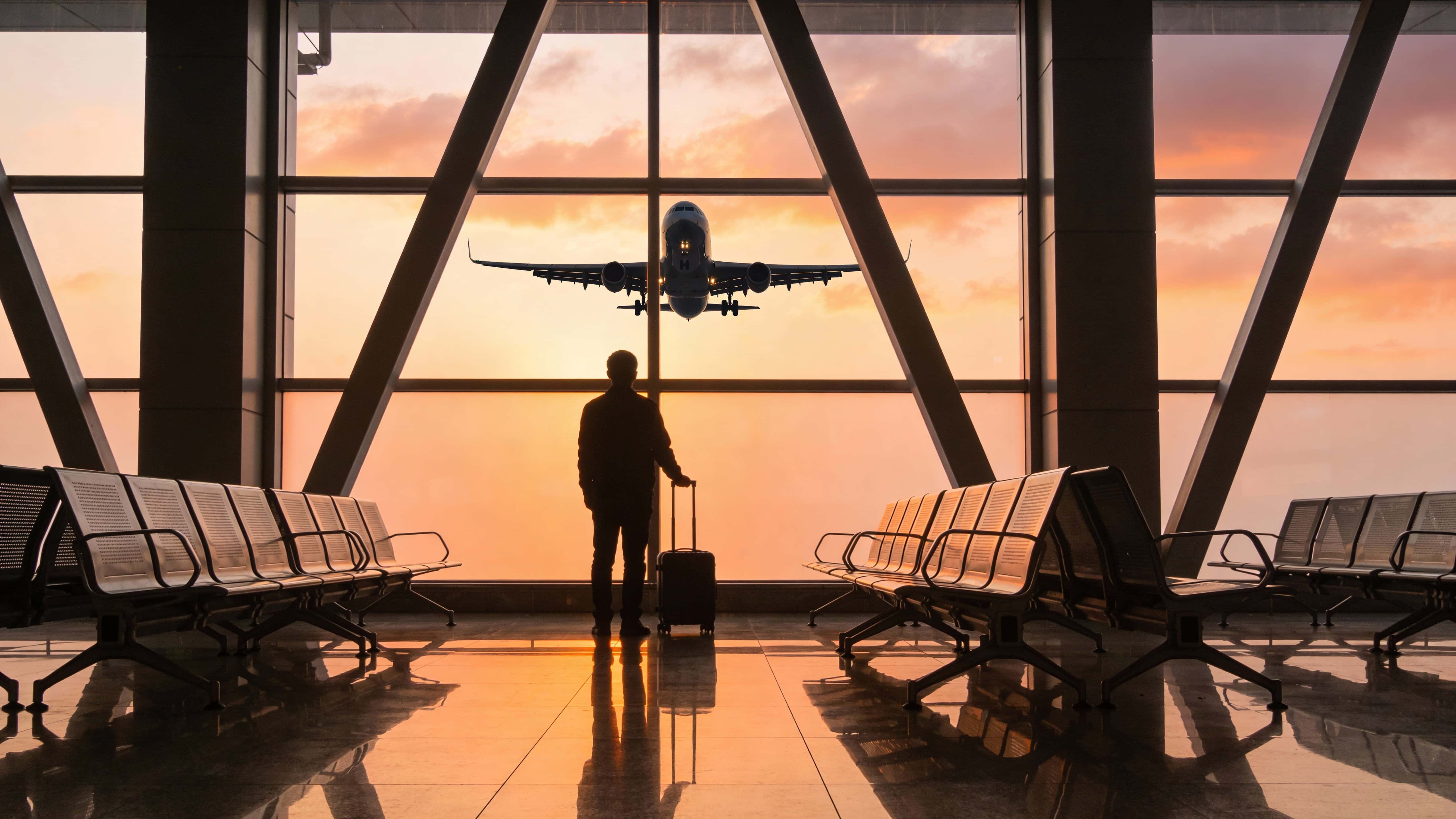A lone traveler stands silhouetted in an airport terminal, holding a small rolling suitcase and facing a large window as an airplane lifts off against a warm orange-peach sunrise.