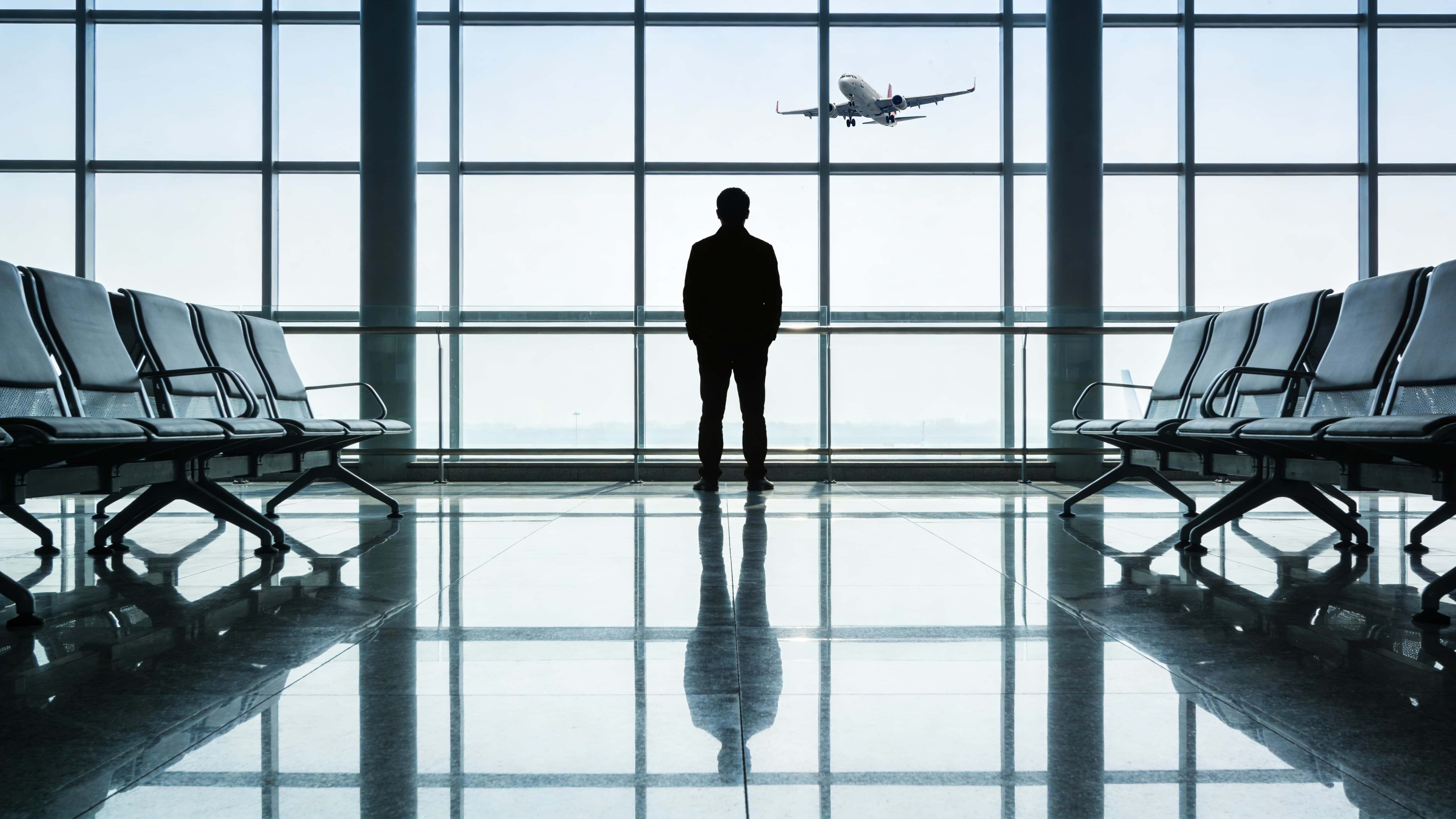 A modern airport interior showing a lone traveler silhouetted against expansive window glass, watching an aircraft glide under a cool gray-blue sky with polished reflective floors.