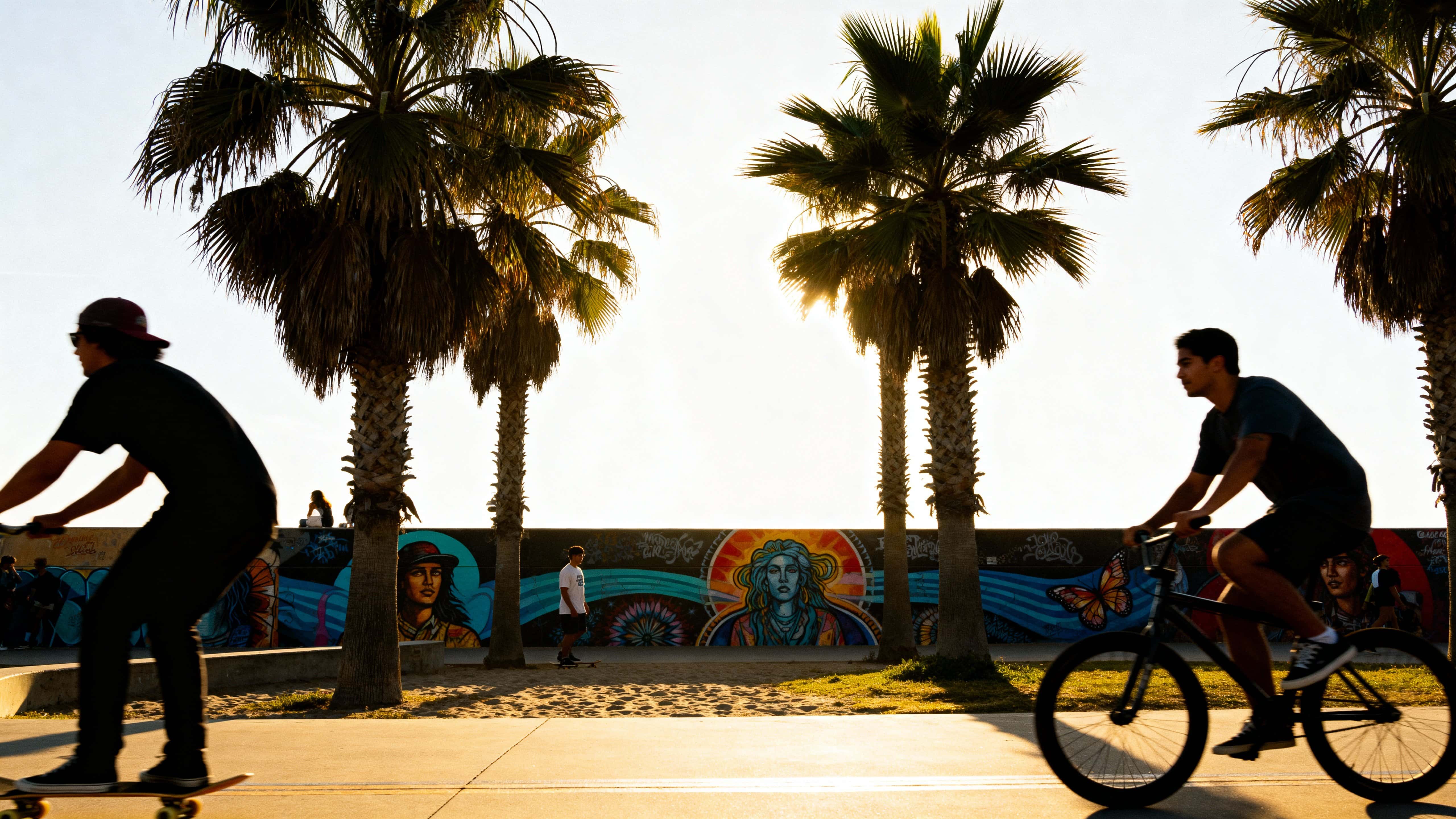 Skaters and cyclists glide along Venice Beach boardwalk beneath swaying palms and vivid murals, bright midday sun, natural reflections, and lively travel energy.