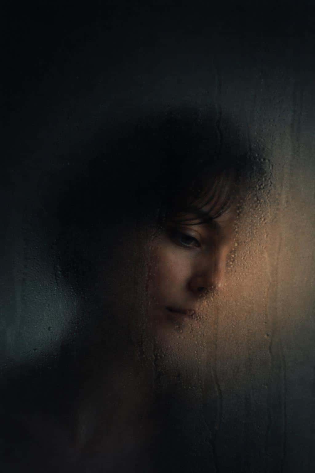 Vertical 2:3 intimate editorial portrait of a woman behind fogged glass, a small cleared patch revealing one eye and cheek while the rest dissolves into soft haze.