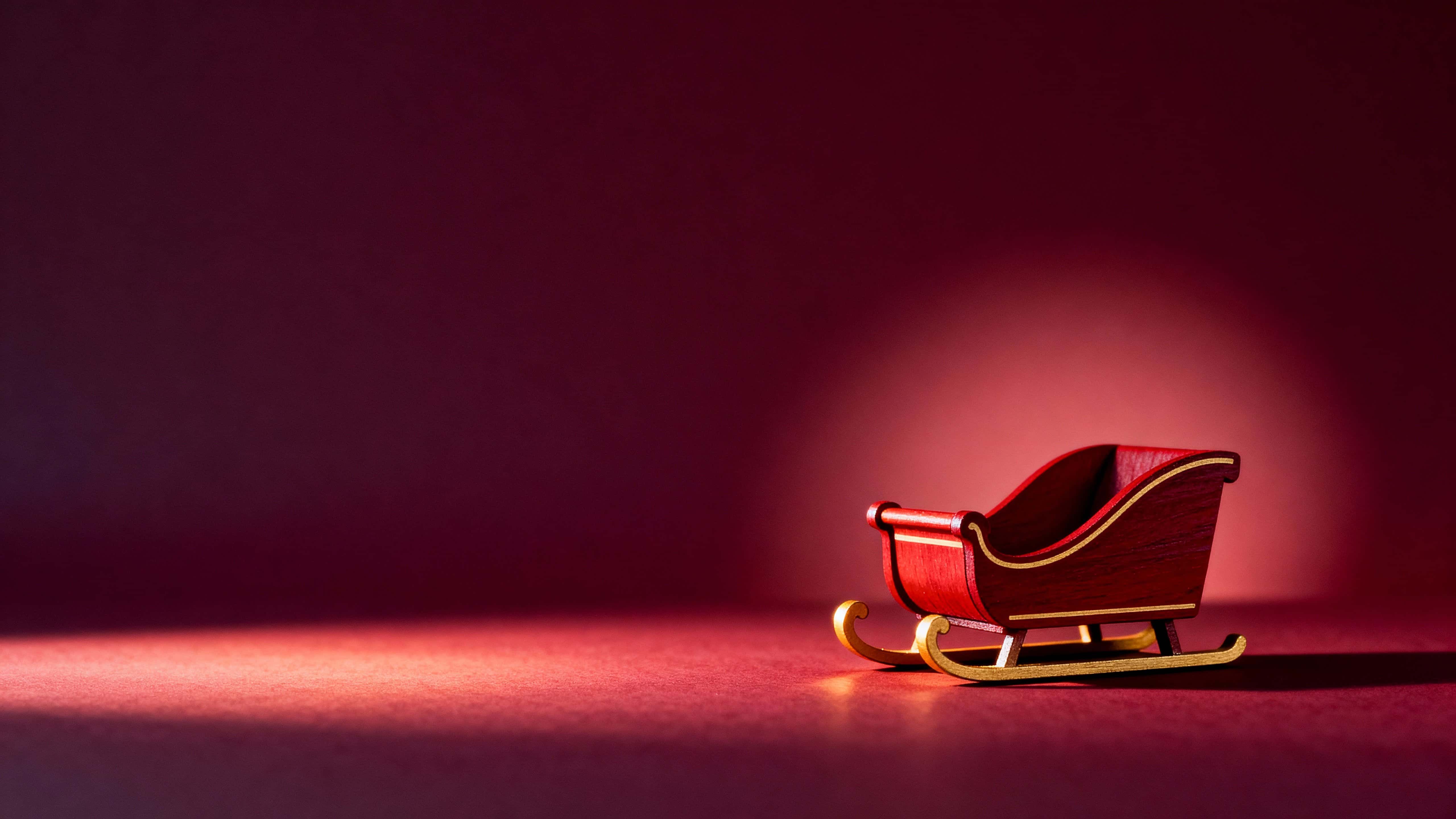 A minimalist festive scene of a small deep-red wooden sleigh with golden runners placed low-right against a gradient wine-red backdrop, lit warmly from the left to create a gentle halo and shadow.