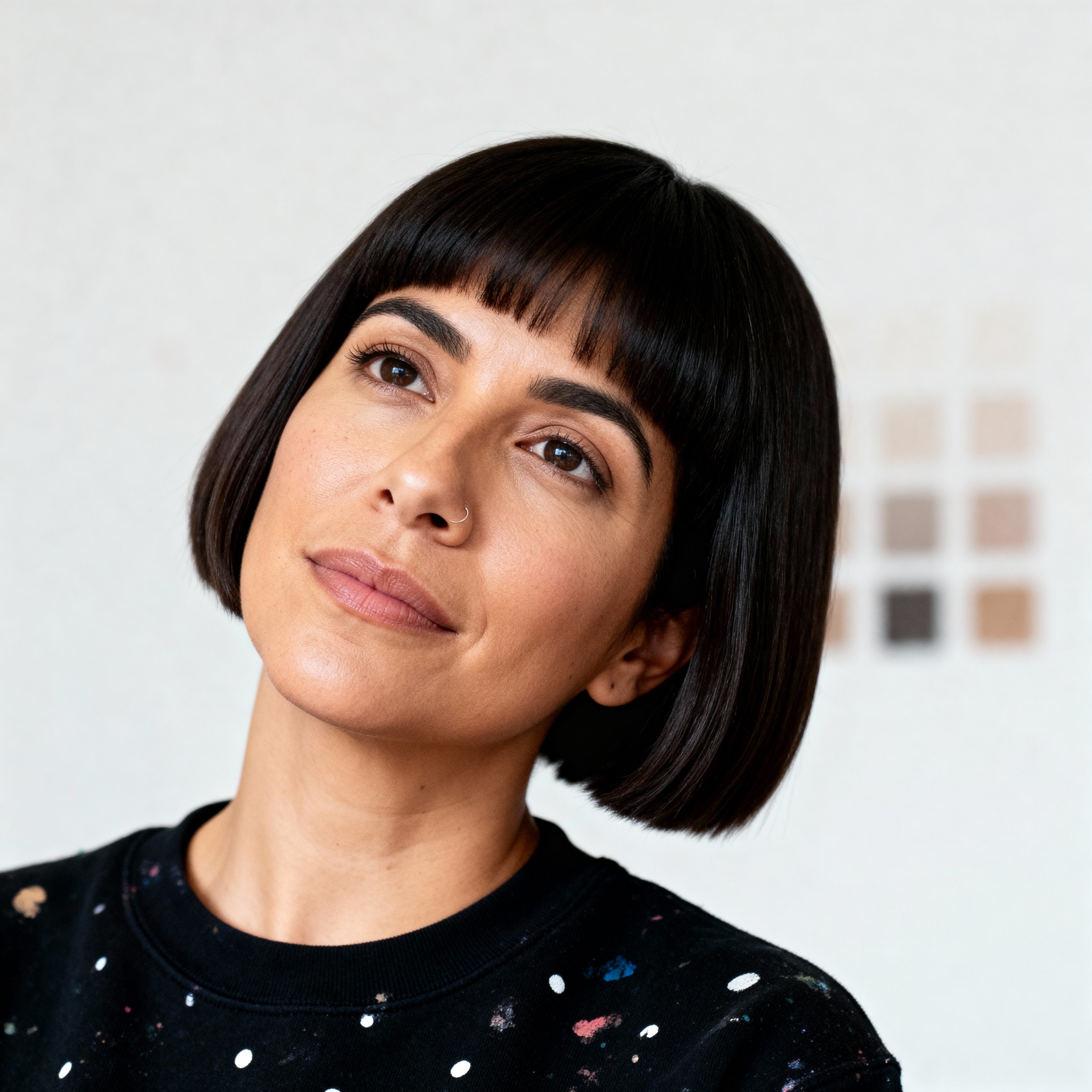 Yareli Cortez, 37, Mexican American set designer in a black crewneck dotted with coral paint specks and a short blunt haircut, tilts her head with steady creative focus under gentle light that smooths tones and lifts cheekbones against a p